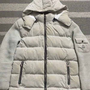 Beige Moncler dunjacka med huva - Säljer en beige Moncler dunjacka med stickade ärmar och huva. Jackan har två dragkedjefickor fram, en ficka med knapp på ärmen och Moncler-logga. Quiltad design, vattenavvisande material och snygga detaljer. Perfekt för kyliga dagar.