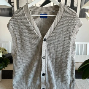 Beige stickad väst från Jack & Jones - Stickad väst från Jack & Jones i beige nyans med grov ribbstickning och svarta knappar framtill. Västen har v-ringning och är tillverkad i 100% bomull, perfekt för lager-på-lager-stil. Enkel och clean design som funkar till många outfits.