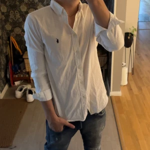 Vit skjorta från Ralph Lauren - Snygg vit skjorta från Ralph Lauren med klassisk krage och blå broderad logga på bröstet. Skjortan har lång ärm och knappar framtill. Perfekt för dig som gillar stilrena och tidlösa plagg med en touch av preppy. Har aldrig använt denhöra bara köpt den på plick 