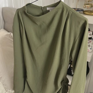 Olivgrön kort klänning från H&M - (har inte kunnat styla den bra därför har jag aldrig haft på mig den) Snygg olivgrön klänning från H&M med lång ärm och rund hals. Klänningen har en cool drapering och knytning i midjan som ger en trendig siluett. Materialet är mjukt och har en lätt glans. Perfekt för dig som gillar minimalistisk stil med en twist.