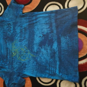 Blå t-shirt med vingar och tryck - Säljer en cool blå t-shirt med korta ärmar och ett unikt mönster i form av stora gula vingar på ryggen. T-shirten har även svart texttryck och detaljer i gult framtill. Materialet känns som mjuk bomull och passformen är normal.