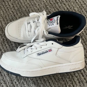 Vita Reebok sneakers i skinn, strl 37 - Klassiska vita Reebok sneakers i skinn med marinblå detaljer och vit sula. Modellen har snörning, perforeringar på tån för extra ventilation och Reebok-logga på sidan och plösen. Snygg och tidlös design som passar till det mesta. Mycket fint skick! Använda några få gånger.