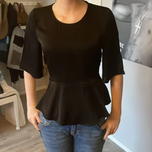 Svart peplumtop från Bik Bok - Säljer en svart peplumtop från Bik Bok med rund halsringning och vida trekvartsärmar. Toppen har en markerad midja och volang nertill samt knäppning bak i nacken och knytband i ryggen. Perfekt att styla med jeans eller kjol.