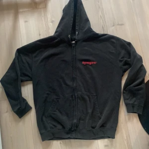 Svart hoodie från Tyngre med dragkedja - Svart hoodie från Tyngre med röd logga på bröstet och ryggen. Tröjan har huva utan snöre (har tappat bort det), hel dragkedja framtill och två stora fickor. Perfekt för gymmet eller chill. Mjuk och skön bomullsblandning med lång ärm. Perfekt i för Vinter-Arc.