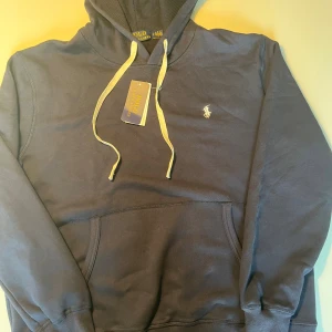 Mörkblå hoodie från Polo Ralph Lauren - Snygg mörkblå hoodie från Polo Ralph Lauren med klassisk broderad logga på bröstet. Tröjan har huva med vita snören, känguruficka och ribbade muddar. Perfekt för en chill och stilren look.