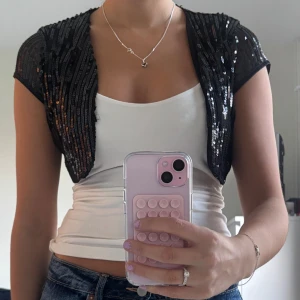 Bolero - Bolero med glittrande paljetter. Västen har korta ärmar och öppen front, täckt av svarta paljetter i vertikala rader. 