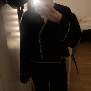 Tun jacka  - Svart tun jacka från Gina Tricot med vita dekorativa sömmar längs kanterna. Koftan har lång ärm och en loose passform, perfekt för lager-på-lager. Enkel och stilren design som ger en cool touch till din outfit.