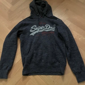Mörkblå Superdry hoodie med tryck - Snygg mörkblå hoodie från Superdry med stort vitt och rött tryck på bröstet. Tröjan har huva med snörning, känguruficka och långa ärmar. Perfekt för dig som gillar en avslappnad streetstil.