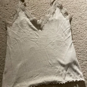 Beige ribbad linnetopp med spets från Amisu - Säljer en beige ribbad linnetopp från Amisu i storlek S. Toppen har smala axelband och v-ringad hals med snygga spetsdetaljer. Materialet är mjukt och stretchigt, perfekt för att skapa en chill och feminin look. Nedre kanten har en lätt vågig finish.
