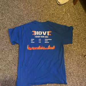 Blå Hov1 tour t-shirt 2024 - Säljer en blå Hov1 4-Ever Tour 2024 t-shirt i storlek M. T-shirten har kort ärm, tryck med turnédatum och eld-detaljer på ryggen samt en liten krona på bröstet. Skön passform och tillverkad i mjuk bomull. Perfekt för dig som älskar Hov1 och vill ha en snygg merch.