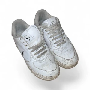 Vita sneakers från Polo Ralph Lauren - Klassiska vita sneakers från Polo Ralph Lauren med låg profil och rund tå. Skorna har snörning, diskret svart logga på sidan och Polo-tryck baktill. Tillverkade i skinn med vit sula och gråvita skosnören. Perfekta för en clean och sportig look.