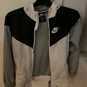 Nike vindjacka loose fit svart/vit - Nike vindjacka i loose fit med svart och vit färgblock-design. Jackan har huva, dragkedja framtill och två sidofickor med dragkedja. Tillverkad i lätt polyester med meshfoder för extra komfort. Perfekt för dig som gillar sportig stil.