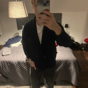 Marinblå cardigan  - Säljer en mörkblå stickad kofta  med dubbla knappar framtill och bred ribbad krage. Koftan har långa ärmar, broderad logga på bröstet och två fickor. Perfekt för dig som gillar klassisk stil med en modern touch.