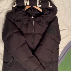 Svart Moncler pufferjacka med huva - Svart pufferjacka från Moncler med stor huva och tydlig logga på ärmen. Jackan har dragkedja framtill och på fickorna, samt quiltad design för extra värme. Perfekt för kalla dagar och har en clean, modern look. Storlek M passar även S. Möts i Stockholm fraktär även
