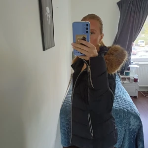 Svart pufferjacka med pälsdetalj - Säljer en svart pufferjacka med fluffig brun fuskpäls runt huvan. Jackan har snygga silverfärgade dragkedjor både framtill och på fickorna. Har ett hå i en av fickorna men de kan man ju fixa. Perfekt för kalla dagar när du vill vara både varm och stilren.