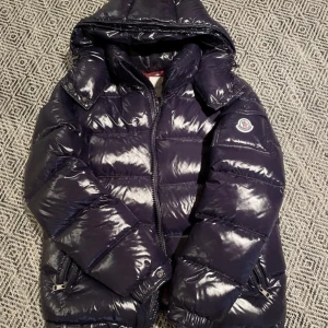 Mörkblå Moncler pufferjacka med huva - Snygg mörkblå pufferjacka från Moncler med glansig finish, huva och dragkedja. Jackan har Moncler-logga på ärmen, flera fickor med dragkedja och quiltad design. Insidan är vinröd med coolt serietryck. Perfekt för kalla dagar när du vill vara både varm och stilren.