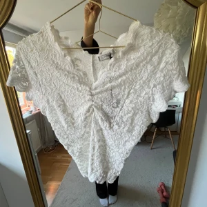 Populär spets topp - Supersöt vit blus från design by si med v-ringning och korta ärmar. Blusen är helt täckt av spets med blommigt mönster och har snygga vågiga kanter. Den har en lätt rynkning framtill som ger en extra touch. Aldrig använd och lappen kvar💕onesize men skulle säga s.