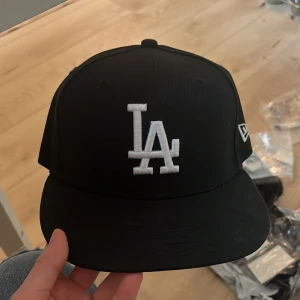 Svart LA keps från New Era - Snygg svart keps från New Era med LA-logga broderad i vitt framtill och MLB-logga baktill. Klassisk flat brim och stilren design, perfekt för dig som gillar streetwear och sportiga vibes. Kepsen är gjord i polyester och har en normal passform. Storlek 56,8cm