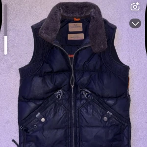  Parajumper väst  - Svart dunväst från Parajumpers med fet pälskrage och coola detaljer som snedställda fickor med dragkedja och tryckknappar. Västen har en grov dragkedja framtill och är fodrad för extra värme. Perfekt för lager-på-lager och streetstyle.