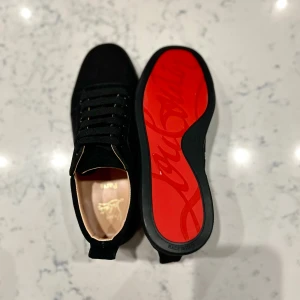 Christian Louboutin svarta sneakers mocka - Säljer ett par svarta sneakers från Christian Louboutin i lyxig mocka med klassisk röd sula och diskret logga undertill. Skorna har rund tå, svarta snören och snygga detaljer i svart. Perfekta för dig som vill sticka ut med exklusiv stil.