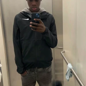 Svart Nike vindjacka med huva - Säljer en svart vindjacka från Nike med huva och hel dragkedja framtill. Jackan har en stilren design med vit Nike-logga på bröstet och är perfekt för dig som gillar sportig streetstyle. Passformen är normal och jackan har långa ärmar.