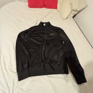 Svart EA7 Emporio Armani tracksuit - Säljer en svart tracksuit från EA7 Emporio Armani med guldfärgad logga på både jacka och byxa. Setet har en stilren design med hel dragkedja på jackan och ribbade muddar på både jacka och byxa. Materialet är mjukt och syntetiskt, perfekt för chill eller träning.
