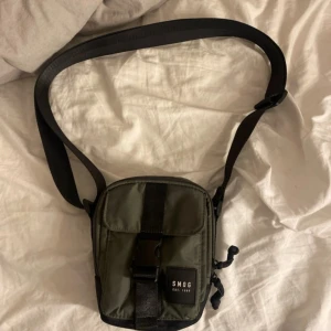 Grön axelväska från SMOG - Snygg grön axelväska från SMOG med svart justerbar rem och praktiskt spänne framtill. Väskan har dragkedja upptill och en extra ficka på framsidan. Perfekt storlek för mobil, plånbok och småprylar. Stilren design med svart detalj längst ner.