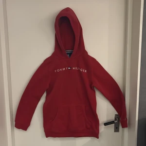 Röd hoodie Tommy Hilfiger XL - Röd hoodie från Tommy Hilfiger i storlek XL. Klassisk modell med huva, magficka och logga tryckt över bröstet. Mjuk insida och ribbade muddar. Perfekt för dig som gillar streetwear och vill ha en snygg och enkel look.