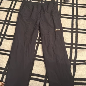 Svarta täckbyxor från Napapijri - Svarta täckbyxor från Napapijri med elastisk midja och snörning. Byxorna har en stilren design med diskret logga på benet och är tillverkade i syntetmaterial med meshfoder. Perfekta för kalla dagar och utomhusaktiviteter.