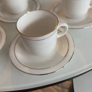 4 vita kaffekoppar med fat – porslin, guldkant, elegant set - Vackert set med fyra vita porslinskoppar med tillhörande fat, dekorerade med en tunn guldkant som ger en elegant och lyxig känsla.  Kopparna är i mycket gott skick – inga nagg, sprickor eller synligt slitage. 