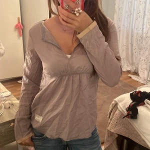 Ljuslila blus från Odd Molly - Supersöt ljuslila blus från Odd Molly med v-ringning och lång ärm. Blusen har en feminin passform med lätt rynk under bysten och beige detaljer vid ärmslut, inklusive en liten rosett. Perfekt för dig som gillar en avslappnad men ändå söt stil.