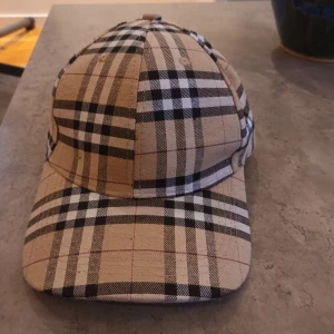 Burberry rutig keps med guldspänne - Snygg keps från Burberry i klassiskt beige, svart och vit rutigt mönster. Kepsen har ett justerbart guldspänne med Burberry-logga och insidan är dekorerad med Burberry-band. Perfekt för dig som vill ha en lyxig och trendig accessoar.