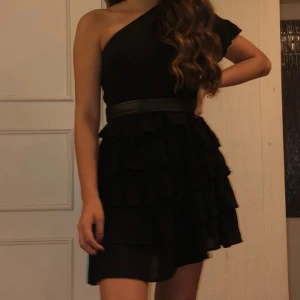 Svart one shoulder volangklänning Boohoo - Svart kort volangklänning från Boohoo med one shoulder-design och flera lager volanger i kjolen. Klänningen är enaxlad. Bandet runt midjan på första bilden tillkommer inte. Storlek 36, normal i storleken. 