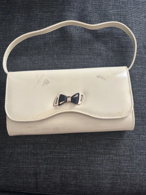 Beige kuvertväska  - Snygg beige kuvertväska i blankt syntetmaterial med guldfärgad kedja och ett dekorativt rosettspänne i svart och guld på framsidan. Väskan har både axelrem och kedja, samt ett innerfack med dragkedja.