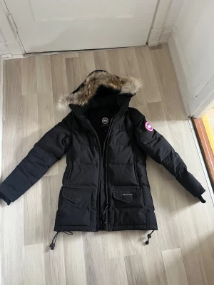Svart dunjacka Canada Goose med päls - Säljer en svart Canada Goose dunjacka med fluffig päls på huvan och klassisk logga på ärmen. Jackan har dragkedja, stora fickor med lock och justerbara snören nedtill. Insidan har praktiska axelband för att bära jackan som ryggsäck. Perfekt för kalla dagar.