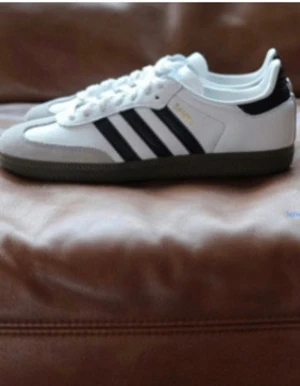 HELT NYA Adidas Samba vita sneakers - Klassiska Adidas Samba sneakers i vitt skinn med svarta ränder och mörkgrå sula. Ikonisk retrostil med snörning och låg profil, perfekt för dig som gillar streetwear och sportig look.