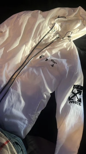 Vit vindjacka från Off-White L - Säljer en clean vit vindjacka från Off-White i storlek L. Jackan har svart dragkedja, huva med snörning och feta logotryck på bröstet och ärmen. Materialet är lätt och syntetiskt, perfekt för att hålla stilen när det blåser. Snygg streetwear-vibe och riktigt schyssta detaljer.