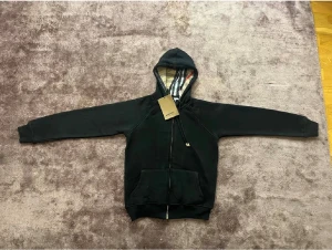 Svart Burberry hoodie med rutig huva - Svart hoodie från Burberry med klassisk rutig insida i huvan och guldfärgade detaljer på snörningen. Tröjan har dragkedja framtill, känguruficka och långa ärmar. Perfekt för dig som vill ha en stilren och lyxig look med ikonisk Burberry-touch.