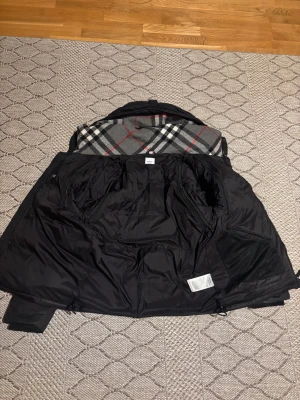 Svart Burberry pufferjacka med huva - Svart pufferjacka från Burberry med huva och klassiskt rutigt foder i grått, vitt och rött. Jackan har tre fickor, där en av de på insidan. Perfekt för kalla dagar och riktigt snygg streetstil.