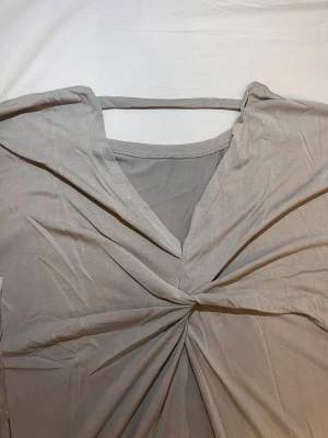 Grå omlott topp med twist fram - Säljer en beige topp från Change Now i storlek L. Toppen har en snygg twistad detalj framtill och omlott-effekt med v-ringning. Den är ärmlös och gjord i mjukt bomullsmaterial, perfekt för en chill och stilren look.