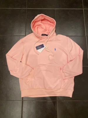 Ljusrosa hoodie från Polo Ralph Lauren - Säljer en ljusrosa hoodie från Polo Ralph Lauren med klassisk huva och snörning. Tröjan har en broderad blå logga på bröstet, känguruficka och ribbade muddar. Tillverkad i mjuk bomull som är skön mot huden. Perfekt för chill dagar.