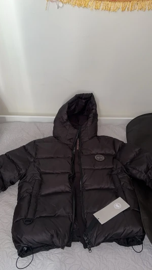 Svart pufferjacka från Canada Goose - Svart pufferjacka från Canada Goose med huva och tydlig logga på bröstet. Jackan har dragkedja framtill, två sidofickor med dragkedja och ett stilrent, quiltat mönster. Perfekt för kalla dagar och har en modern, oversized look. Den är i m men passar L. Och är använd lite grann 