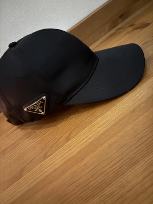 Svart keps från Prada i nylon - Säljer en svart keps från Prada med klassisk böjd skärm och justerbar rem bak. Kepsen har Pradas ikoniska metallogo på sidan och är tillverkad i glansig nylon för en clean och lyxig vibe. Perfekt accessoar för att lyfta din streetstyle.