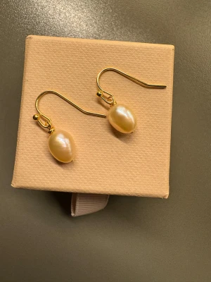 Guldörhängen med sötvatten pärlor  - Snygga örhängen med droppformade sötvatten pärlor i ca strlk 5mm  och guldfärgade krokar. Pärlorna har en naturlig glans och hänger elegant från örat. Perfekt för dig som gillar klassisk och stilren design med en touch av lyx.