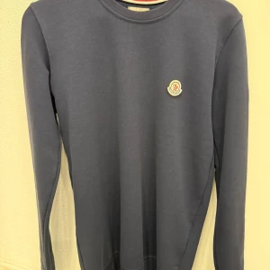 Mörkblå långärmad tröja Moncler - Snygg mörkblå långärmad tröja från Moncler med rund hals och diskret logotyp på bröstet. Tröjan har en clean look och är tillverkad i mjuk bomull som känns skön mot huden. Perfekt för dig som gillar stilrena plagg med exklusiv känsla.