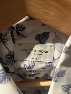Vit blommig skjorta Bruun & Stengade - Snygg vit skjorta från Bruun & Stengade med blått blommigt mönster. Slim fit-modell med långa ärmar och klassisk krage. Skjortan är tillverkad i bomull och har knappar framtill. Perfekt för dig som gillar stilrena detaljer och easy care-material.
