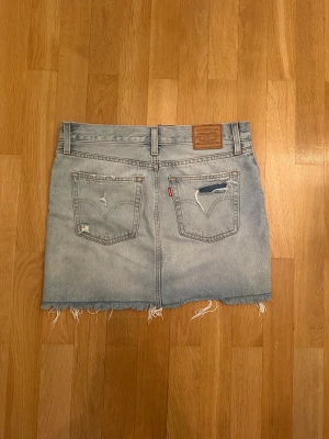 Levi's ljusblå jeanskjol  - Säljer en ljusblå jeanskjol från Levi's i klassisk modell med slitna detaljer och fransig nederkant. Kjolen har knappstängning framtill. Storlek 28! 
