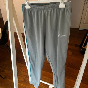 Grå träningsbyxor Nike x Joan Bonito - Snygga grå träningsbyxor med vita meshpaneler på sidorna och turkosa detaljer längs benen. Nike-logga på framsidan och Joan Bonito-märke vid fickan. Byxorna har elastisk midja och dragkedjefickor, perfekta för sport eller chill.