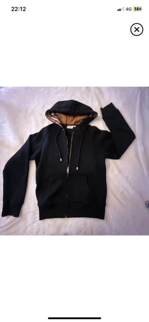Svart Burberry hoodie med dragkedja - Svart hoodie från Burberry med klassiskt rutigt foder i huvan. Tröjan har dragkedja framtill, snörning vid huvan och två fickor. Materialet är mjuk bomull och passformen är normal. Perfekt för dig som gillar stilrena och lyxiga streetwear-plagg.