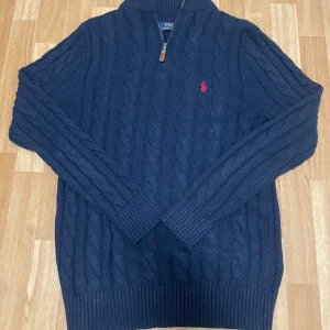 Marinblå stickad tröja  - Klassisk och stilren mörkblå kabelstickad tröja läderdetalj på dragkedjan. Röd broderad logga på bröstet. Perfekt både till vardags och lite mer uppklätt.  Skick: Mycket bra begagnat skick – inga fläckar eller hål, endast lite normalt ludd. Storlek: (L.) Passform: Regular fit Färg: Mörkblå (navy)  Superfin kvalitetsstickad tröja som håller länge.